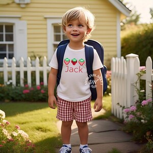 May include: A child in a white t-shirt with strawberry designs and the name "Jack". They wear red and white checked shorts and a blue backpack. The background shows a yellow house and a white picket fence, typical of a suburban setting.