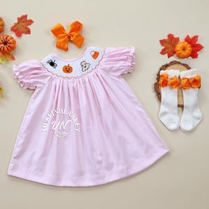 May include: A pink dress with a Peter Pan collar and embroidered Halloween designs, including a pumpkin, ghost, spider, and candy corn. The dress has short puffed sleeves and a gathered skirt. The dress is paired with white knee-high socks with orange bows.