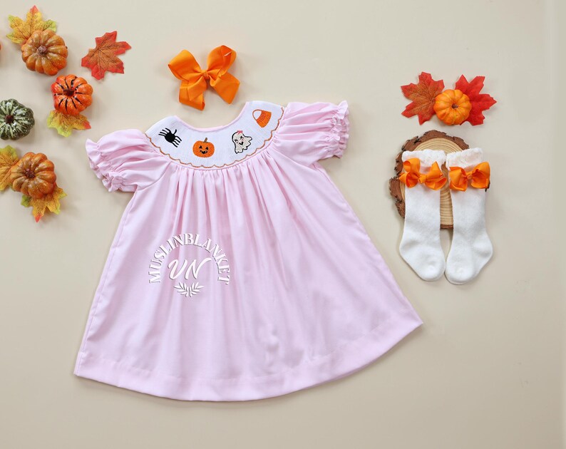 May include: A pink dress with a Peter Pan collar and embroidered Halloween designs, including a pumpkin, ghost, spider, and candy corn. The dress has short puffed sleeves and a gathered skirt. The dress is paired with white knee-high socks with orange bows.