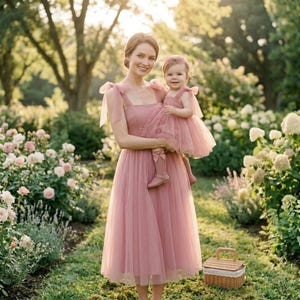 May include: A woman and a child wearing matching dusty rose tulle dresses. The woman is holding the child in a garden setting with a picnic basket. The dresses have bow details on the shoulders and the child is wearing matching socks.