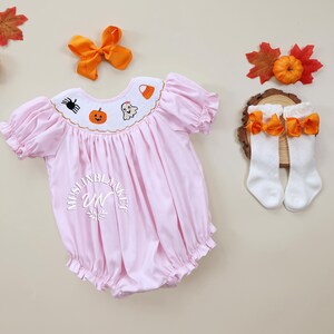 May include: A pink baby onesie with embroidered Halloween designs, including a pumpkin, ghost, spider, and candy corn. The onesie has a ruffled collar and short sleeves. The onesie is paired with white socks with orange bows.
