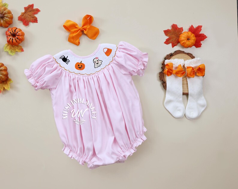 May include: A pink baby onesie with embroidered Halloween designs, including a pumpkin, ghost, spider, and candy corn. The onesie has a ruffled collar and short sleeves. The onesie is paired with white socks with orange bows.