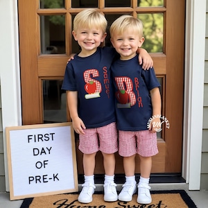 May include: Two children in navy t-shirts with apple and name designs, and red and white gingham shorts. They stand before a wooden door, with a sign saying "FIRST DAY OF PRE-K". The image captures a back-to-school theme.