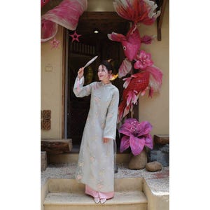 May include: A woman in a pale green, long-sleeved dress with floral embroidery, holding a fan. The dress has white cuffs and a pink hem. Decorative paper fish and flowers decorate the background, creating a festive atmosphere.