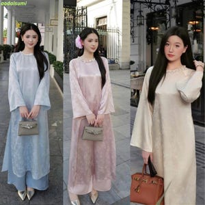 Puede incluir: Tres mujeres con vestidos tradicionales vietnamitas Áo dài en diferentes colores: azul claro, rosa y crema. Cada vestido tiene mangas largas, cuello alto y un estampado floral. Sostienen pequeños bolsos.