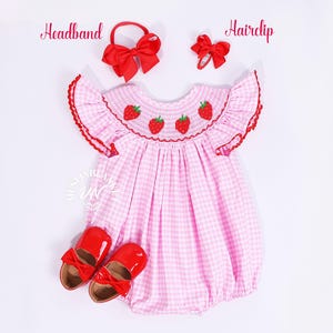 May include: Pink and white gingham baby dress with embroidered strawberries. The dress has puffed sleeves and a smocked bodice. It comes with a red headband and a red hair clip.