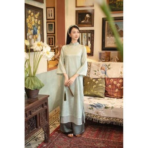 Puede incluir: Un vestido verde claro de manga larga con cuello alto, usado con pantalones de pernera ancha. El vestido tiene una abertura en el dobladillo. La mujer sostiene un abanico decorativo. El fondo incluye obras de arte enmarcadas y un sofá floral.