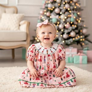 May include: A baby wearing a pink dress with a nutcracker pattern and a pink bow headband. The dress has a white collar and green trim. A Christmas tree with baubles is in the background.