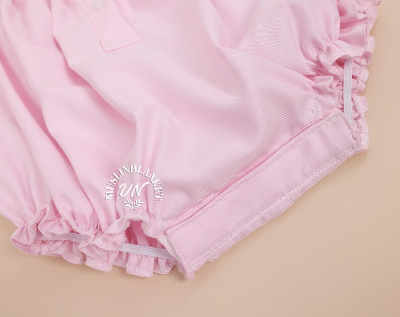 May include: Pink baby bloomers with a ruffled edge and elastic waistband. The bloomers have a white elastic band around the leg openings.