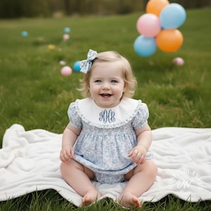 Puede incluir: Un bebé sonriente con un pelele floral azul con cuello blanco y lazo a juego. El cuello tiene iniciales bordadas. El bebé está sentado en una manta blanca sobre un campo de hierba, con globos de colores al fondo.