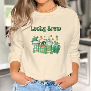 Puede incluir: Una sudadera de color crema con el texto "Lucky Brew" impreso en la parte delantera. El diseño presenta una ilustración de dibujos animados de tres bebidas verdes y doradas con tréboles y monedas de oro.