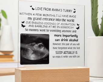Op de afbeelding: Een transparant acryl frame met een zwart-witte echografie afbeelding van een baby. Het frame heeft een houten basis en bevat een boodschap over de vreugde van het ouderschap, inclusief de mogelijkheid om weer alcohol te drinken. De tekst luidt: "LOVE FROM MAMA'S TUMMY WITHIN A FEW MONTHS, I'LL HAVE MADE MY GRAND ENTRANCE INTO THE WORLD CUE ENDLESS COOINGS OF ADORATION AND BABBLING AT ME IN GIBBERISH So mommy wont be vomitting anymore and more importantly can drink alcohol however, the pair of you will have forgotten what the hell SLEEP ACTUALLY IS so enjoy it while you still can."