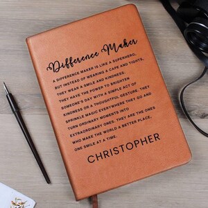 Könnte beinhalten: Ein braunes Lederjournal mit dem Text "Difference Maker" und einem Zitat darüber, wie man einen Unterschied in der Welt macht. Das Journal ist mit dem Namen "Christopher" personalisiert.