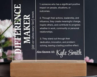Pode incluir: Placa de acrílico transparente com a palavra "DIFFERENCE MAKER" em branco, montada em uma base de madeira. A placa inclui uma definição e o nome "Kyle Smith". O fundo é escuro com detalhes florais.