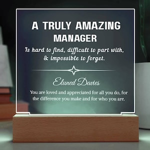 Può includere: Targa luminosa in acrilico con il testo "A Truly Amazing Manager" e un messaggio personalizzato. La targa è montata su una base in legno, ideale per una scrivania o uno scaffale. Il testo è bianco su sfondo scuro.