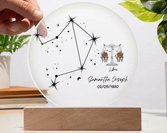 May include: A white round acrylic sign with a black Libra zodiac constellation design, a floral Libra symbol, and the text "Samantha Joseph 02/05/1990" and "Libra". The sign is on a wooden base.