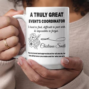 May include: White ceramic mug with black text that reads "A Truly Great Events Coordinator." The mug also features a floral design and the name "Chistiana Smith." The mug is held by a person.