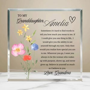 May include: A clear glass block with a floral design and text. The text reads "To My Granddaughter, Amelia" with a heartfelt message from "Love, Grandma." The floral arrangement features pink, yellow, blue, and red watercolor flowers.