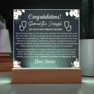 May include: A graduation gift featuring a clear acrylic panel with the text "Congratulations! Samantha Joseph ON YOUR DOCTORATE DEGREE" and a message. The panel is adorned with floral accents and a stethoscope graphic, set on a wooden base.