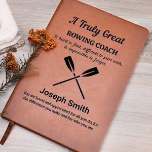 Puede incluir: Un diario de cuero marrón con el texto "A Truly Great Rowing Coach" y el nombre "Joseph Smith". El diario presenta remos cruzados y una cita de agradecimiento. Una ramita de flores naranjas descansa a un lado.