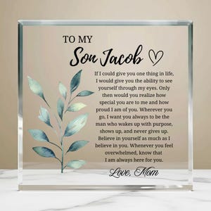 Peut inclure: Un bloc de verre transparent avec le texte "TO MY Son Jacob" et un message de "Maman". Le bloc présente un motif de feuilles de style aquarelle en bleu et vert, idéal comme cadeau.
