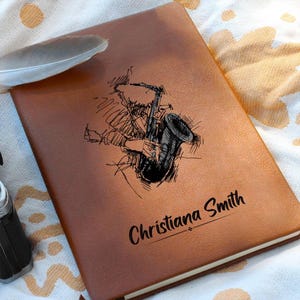 Puede incluir: Un diario de cuero marrón con un boceto de una persona tocando el saxofón. El diario tiene el nombre "Christiana Smith" grabado en la portada.