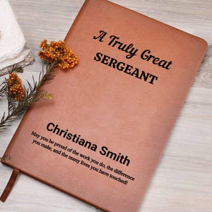 May include: A brown leather journal with the text "A Truly Great SERGEANT" and the name "Christiana Smith" printed in black. The journal includes an inspirational quote. A sprig of orange flowers rests on the journal.
