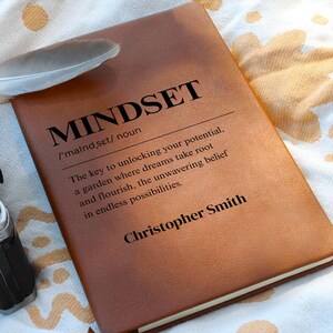 Peut inclure: Un journal en cuir marron avec le mot "MINDSET" imprimé en noir. La définition de l'état d'esprit est imprimée en dessous, ainsi que le nom "Christopher Smith". Une plume repose sur le journal.