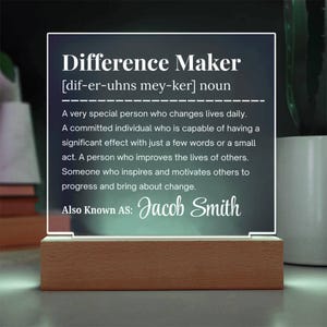 May include: A lit acrylic sign with the words "Difference Maker" and a definition. The sign is mounted on a wooden base. The definition describes a person who changes lives and inspires others. The name "Jacob Smith" is also displayed.