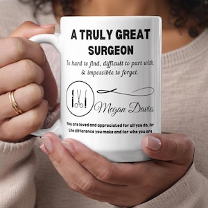 May include: White ceramic mug with the text "A Truly Great Surgeon" in black. The mug features a graphic of surgical tools and the name "Megan Davies". The mug also has a quote about surgeons.