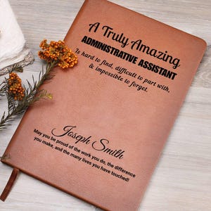 May include: A brown leather journal with the text "A Truly Amazing ADMINISTRATIVE ASSISTANT" and a personalized name. The journal has a ribbon bookmark and is decorated with a sprig of orange flowers.