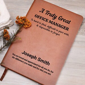 May include: A brown leather journal with the text "A Truly Great OFFICE MANAGER" and the name "Joseph Smith". The journal has a ribbon bookmark and is decorated with a sprig of orange flowers.
