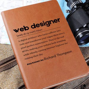 May include: A brown leather-bound journal with the definition of "web designer" printed on the cover. The definition reads: "A digital architect who turns caffeine into code and transforms your vague ideas into a pixel-perfect masterpiece. They're the real MVPs, making your website look stunning while silently cursing Internet Explorer for ruining their day." The text also includes "Also Known As: Richard Thompson."