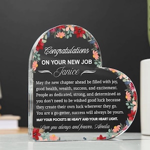Puede incluir: Placa acrílica en forma de corazón con detalles florales. La placa presenta las palabras "Felicitaciones por tu nuevo trabajo" y un mensaje personalizado. Los bordes están adornados con flores rojas, rosas y blancas, creando un borde decorativo.