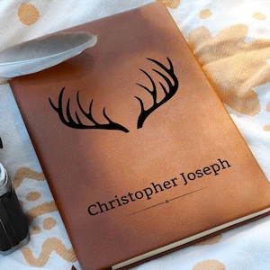 Puede incluir: Un diario de cuero marrón con un diseño de cuernos negros y el nombre "Christopher Joseph" impreso en la portada. Una pluma blanca descansa sobre el diario. El diario está cerrado y tiene una superficie texturizada.