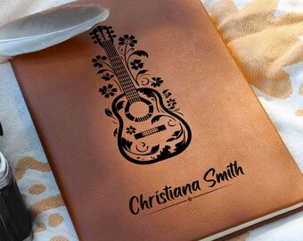 May include: A brown leather journal with a black guitar and floral design. The name "Christiana Smith" is printed in cursive script at the bottom. A feather rests on the journal's cover.
