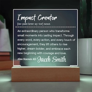 以下が含まれることがあります： 「Impact Creator」という言葉とその定義が書かれた、光るアクリル製のサイン。木製の台座に取り付けられています。テキストには「Jacob Smith」という名前が含まれています。
