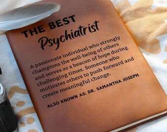 May include: A brown leather-bound journal with the words "THE BEST Psychiatrist" embossed on the cover. The text below describes a passionate individual. A white feather rests on the journal.