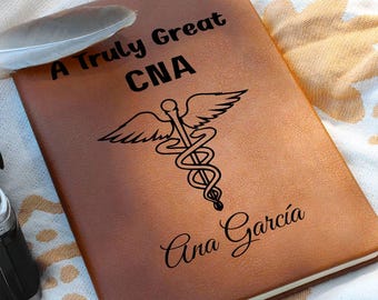 May include: A brown leather-bound notebook with the text "A Truly Great CNA" and the medical caduceus symbol. The name "Ana Garcia" is printed below the symbol. A feather rests on the top left corner.
