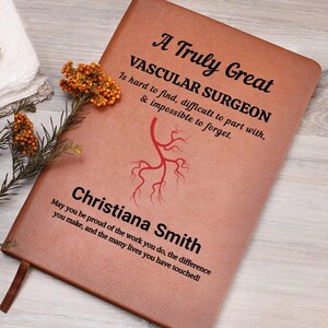 Puede incluir: Un diario de cuero marrón con el texto "A Truly Great Vascular Surgeon" y una ilustración vascular roja. El diario está personalizado con el nombre "Christiana Smith" y un mensaje de apoyo. Una cinta de marcador marrón es visible.