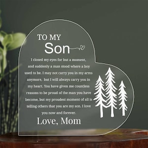 Könnte beinhalten: Herzförmige transparente Acrylplakette mit dem Text "TO MY Son" und einer Botschaft von "Love, Mom". Das Design beinhaltet weiße Baumsilhouetten, geeignet als Geschenk.