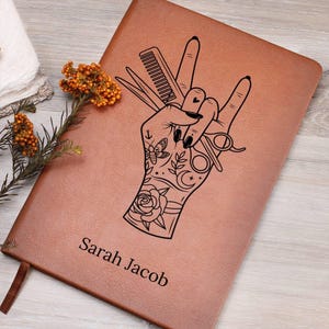 May include: A brown leather journal with a black line drawing of a hand holding scissors and a comb. The hand is tattooed with a rose, a butterfly, a crescent moon, and an anchor. The text "Sarah Jacob" is printed on the front of the journal.