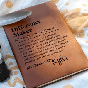 Pode incluir: Um diário de couro castanho com as palavras "Difference Maker" e uma definição impressa em preto. O diário também inclui o nome "Kyler" e uma pena.