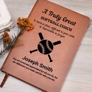 Op de afbeelding: Een bruin leren notitieboek met de tekst "A Truly Great SOFTBALL COACH" en een softbalafbeelding. Het notitieboek bevat de naam "Joseph Smith" en een persoonlijke boodschap. Een takje met oranje bloemen ligt ernaast.