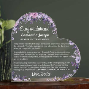 May include: Heart-shaped acrylic plaque with floral accents and the words "Congratulations! Samantha Joseph ON YOUR DOCTORATE DEGREE." The plaque includes an inspirational message and is a graduation gift.