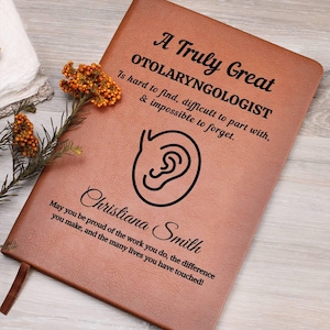 May include: A brown leather journal with the text "A Truly Great OTOLARYNGOLOGIST" and an ear graphic. The journal includes the name "Christiana Smith" and a heartfelt message. A decorative sprig of orange flowers is placed near the journal.