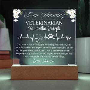 May include: A personalized LED light-up sign for a veterinarian. The clear acrylic panel features the text "To an Amazing Veterinarian" and a name, with a heartbeat line, stethoscope, and paw print design. It rests on a wooden base.