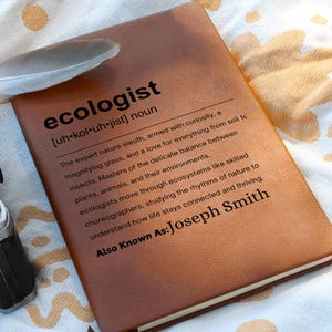 Può includere: Un taccuino marrone rilegato in pelle con la parola "ecologist" stampata in nero. La definizione di ecologista è stampata sotto la parola. Il taccuino è personalizzato con il nome "Joseph Smith". Una piuma è appoggiata nell'angolo in alto a sinistra.
