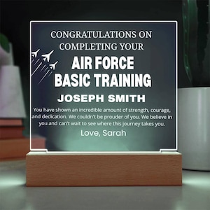 May include: A rectangular illuminated plaque with a wooden base. The plaque displays the text "CONGRATULATIONS ON COMPLETING YOUR AIR FORCE BASIC TRAINING JOSEPH SMITH" in white. The design includes stylized jet illustrations and a personal message.