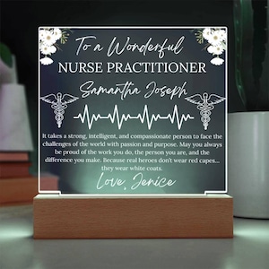 Puede incluir: Luz de noche LED personalizada con base de madera. El panel acrílico transparente presenta el texto "To a Wonderful Nurse Practitioner" y un nombre, junto con una línea de latidos y un símbolo médico. Adornos florales blancos en la parte superior.